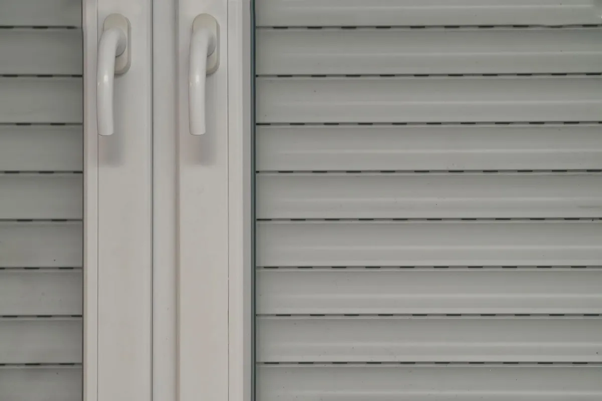 Tablier de volet roulant blanc vu de l'intérieur derrière une fenêtre PVC à deux poignées